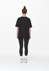 Černé oversized tričko s krátkým rukávem, spárované s černými legínami a bílými ponožkami. Modelka v bílých sportovních botách, pohled zezadu.