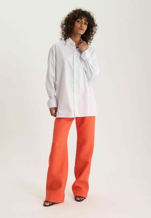 Femme aux cheveux bouclés portant une chemise blanche oversize boutonnée et un pantalon large orange vif, debout sur un fond clair.