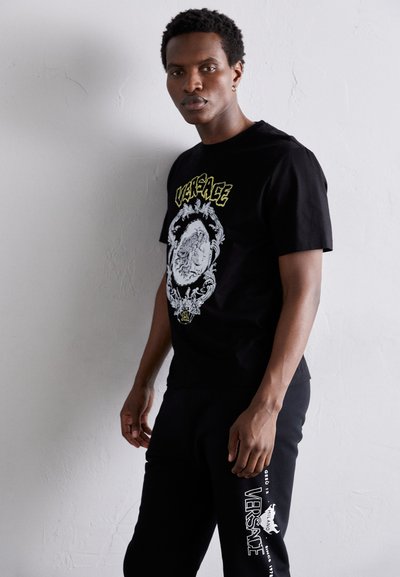 Must tee-shirt musta musta valge ja roheline lõviga ja tekstiga "VERSACE"; kokku sobitatud mustade dressipükstega, millel on "VERSACE" logo.
