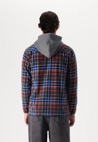 Geruite flanellen blouse met een grijze capuchon, in een rood, blauw en zwart ruitpatroon. Lange mouwen en een ontspannen pasvorm. Zachte textuur.