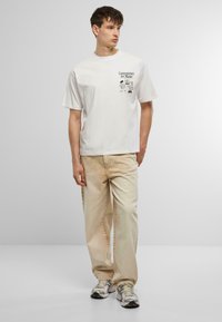 Witte katoenen t-shirt met korte mouwen, voorzien van een zwart grafisch ontwerp. Combinatie met lichtbruine spijkerbroek en grijze sportschoenen.