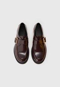 Kliendi tumedast pruunist läikivast nahast monk strap kingad kuldsete pandlitega, mustade talladega ning siseselt on kiri "BOSS", pildistatud valgel taustal.