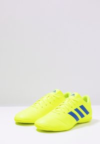 Scarpe da calcio gialle brillanti con una superficie texture, dotate di tre strisce blu e di un logo blu sul tallone, progettate per il turf.