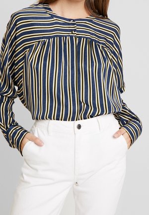 Gestreifte Bluse in Marineblau und Gelb mit rundem Ausschnitt und Knopfdetail, kombiniert mit weißen hochtaillierten Hosen. Weicher Stoff mit lockerer Passform.