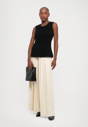 Donna che indossa un top nero senza maniche in pelliccia, pantaloni larghi crema, tiene in mano una borsa nera, in piedi davanti a uno sfondo bianco semplice.