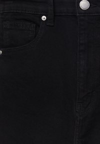 Gros plan sur un jean en denim noir montrant la poche avant, la boucle de ceinture et le bouton argenté avec gravure de la marque.
