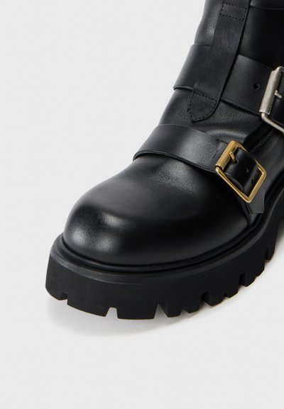 Bota de cuero negra con una punta redondeada, que presenta una correa ancha y un detalle de hebilla, sobre una suela de goma gruesa con un patrón de relieve texturizado.