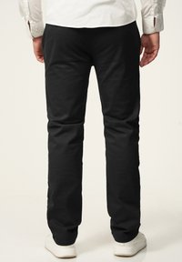 Schwarze Baumwollhose mit schmaler Passform, mit glatter Textur und schlichter Gestaltung. Tragen mit weißen Sneakers, zeigt die Rückansicht.