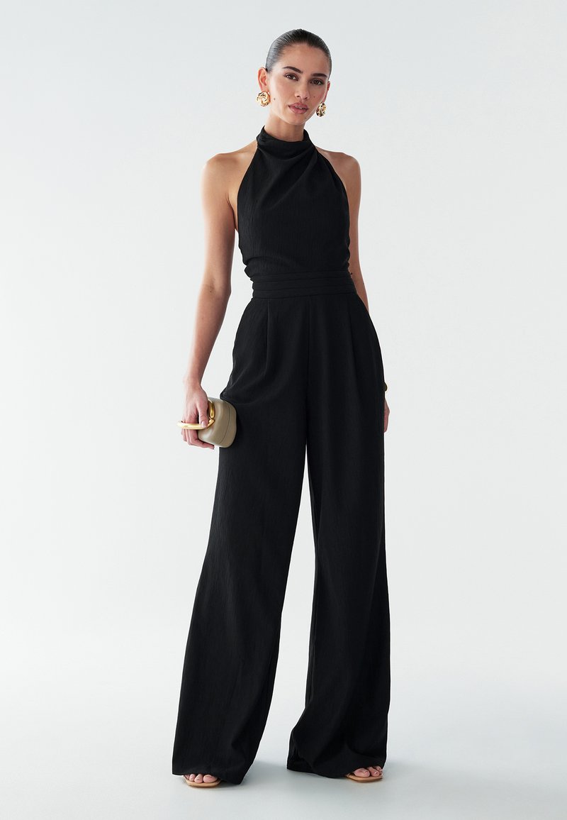 Melna halter stila kombinezons ar pieguļošu augšdaļu un platām biksēm. Tekstūrēta auduma, kas izceļ gludu siluetu un minimālistisku dizainu.