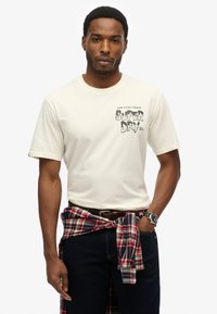 Crème kleurige t-shirt met korte mouwen en een zwart grafisch ontwerp, gecombineerd met donkere jeans en een rode geblokte overhemd dat om de taille is geknoopt. Geaccentueerd met een horloge.