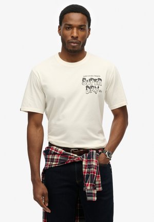 T-shirt crème à manches courtes avec un imprimé graphique noir, associé à un jean foncé et une chemise à carreaux rouge nouée à la taille. Accentué par une montre.