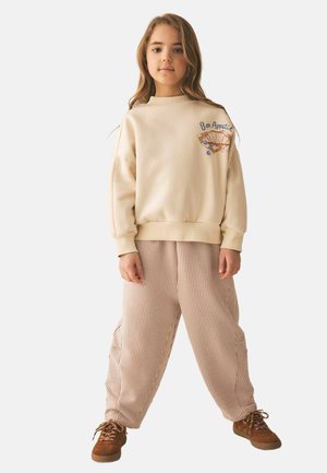 Ung pige står med hænderne ved siden af kroppen iført en beige sweater med "Bon Appetit" og mønstrede bukser med brune sko.