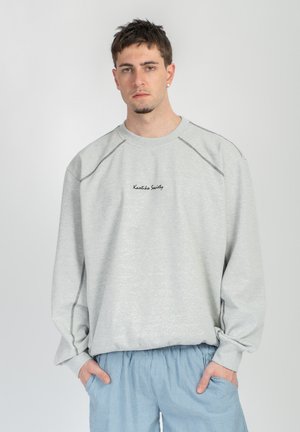 Ung mand med kort hår iført en lysegrå sweatshirt med sort tekst og blå stribede bukser, hænderne i lommerne, foran en ensfarvet baggrund.