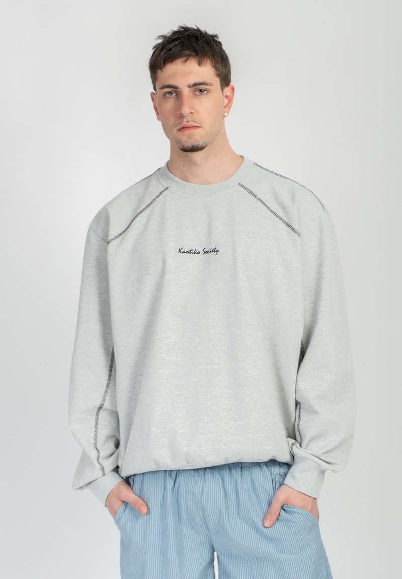 Νεαρός με κοντά μαλλιά φοράει ανοιχτό γκρι φούτερ με μαύρο κείμενο και μπλε ριγέ παντελόνι, με τα χέρια στις τσέπες, μπροστά σε απλό φόντο.