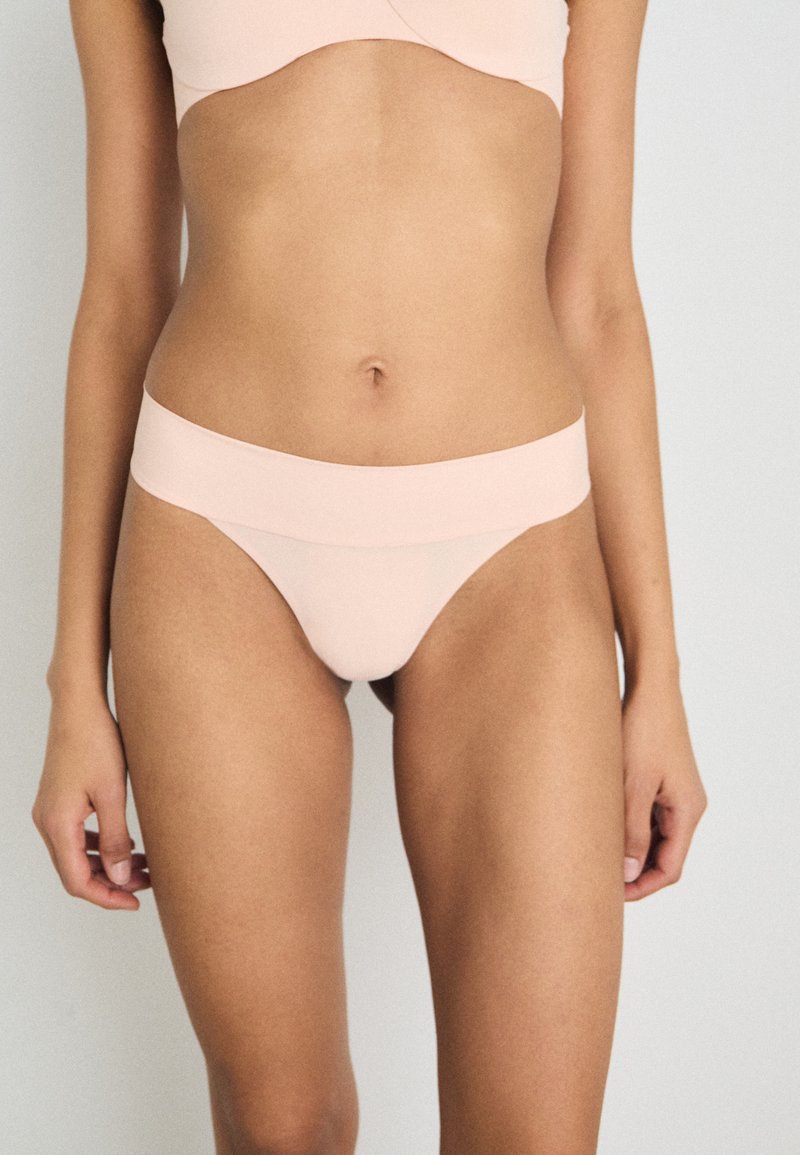 String léger rose, sans couture, fabriqué en tissu lisse avec une large ceinture. Il présente un design minimaliste qui épouse le corps.