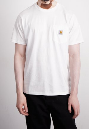 Homme portant un t-shirt blanc uni à manches courtes avec une petite poche et un logo sur le côté gauche de la poitrine, assorti à un pantalon noir.