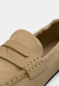 Μπεζ σουέντ μοκασίνι με ραμμένη λεπτομέρεια, απαλό υλικό και ανυψωμένη, στρογγυλεμένη γλώσσα. Η σόλα είναι επίπεδη με ελάχιστες λεπτομέρειες.