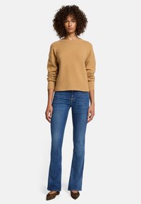 Camel-kleurige trui met ribgebreide zoom, gecombineerd met blauwe flared jeans. De drager staat op een effen achtergrond, waardoor de pasvorm en stijl van de outfit goed zichtbaar zijn.