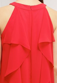 Top sans manches rouge avec un décolleté rond, des volants superposés dans le dos et une fermeture éclair discrète au niveau du cou.