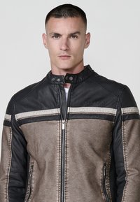 Giovane uomo con capelli corti che indossa una giacca di pelle zip nera e beige con colletto a scatto su uno sfondo semplice.