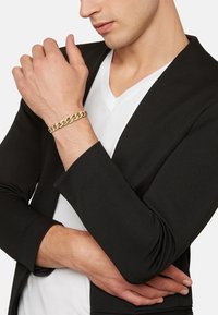 Bracciale in oro con maglie intrecciate indossato al polso, abbinato a una giacca nera e a una maglietta bianca a scollatura a V. Finitura in metallo liscia.