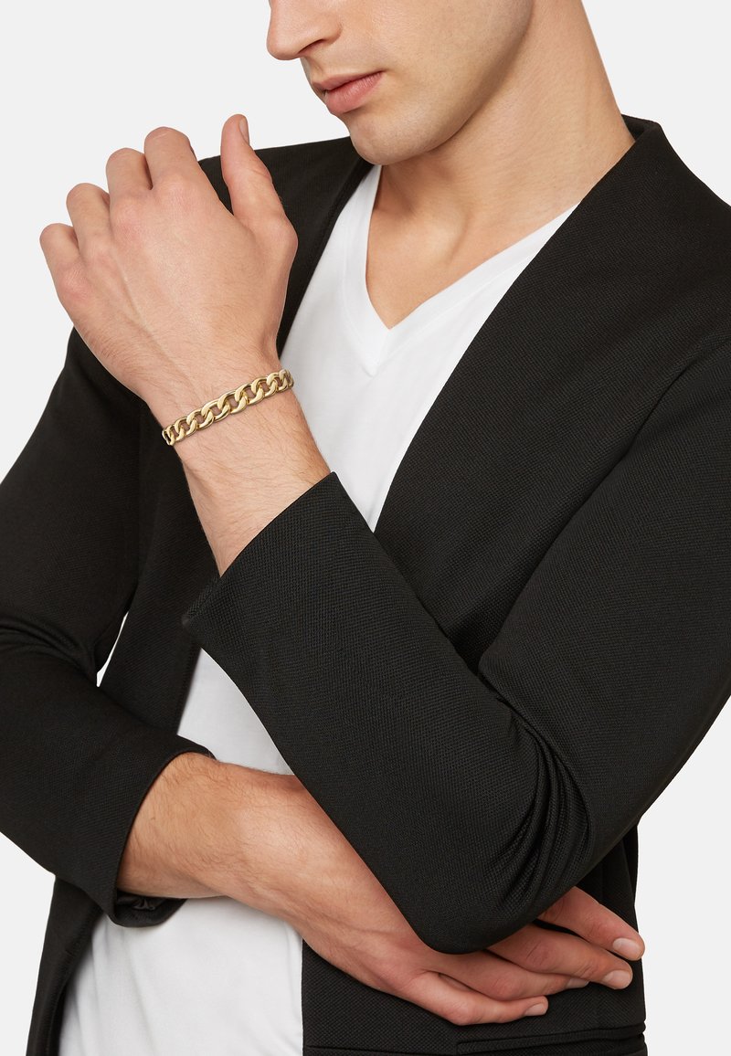 Bracciale in oro con maglie intrecciate indossato al polso, abbinato a una giacca nera e a una maglietta bianca a scollatura a V. Finitura in metallo liscia.