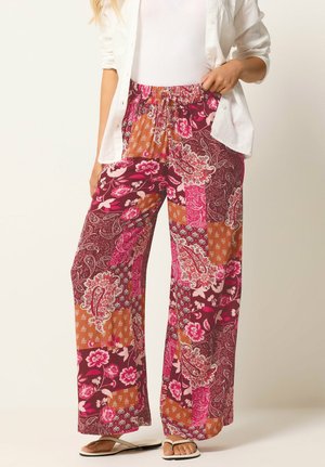 Vrouw draagt wijde, patchwork bloemenbroek in roze, paars en oranje tinten, gecombineerd met witte sandalen en een wit overhemd open gedragen over een wit topje.