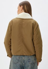 Brun bomberjakke med en hvid shearlingkrave, der har et cropped design, lange ærmer og elastisk talje. Bæres med blå denim jeans.