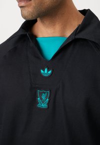 Sort fodboldtrøje med spids krave, turkise detaljer, broderet Adidas-logo og Liverpool FC-crest placeret i centrum.