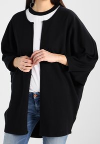 Svart oversized kofta med rund halsringning, öppen front och lösa ärmar, tillverkad av ett mjukt, texturerat tyg. Minimalistisk design.