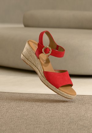 Sandale à talon compensé rouge avec bride cheville, boucle dorée et semelle en espadrille tressée, posée sur un tapis beige texturé devant un canapé beige.