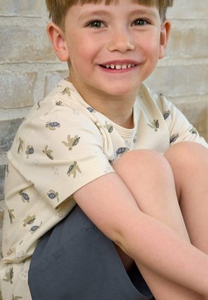 Lächelnder Junge, der mit hochgezogenen Knien sitzt, trägt ein cremefarbenes Hemd mit Schildkröten-Motiven und dunkle Shorts vor einem leicht strukturierten Hintergrund.