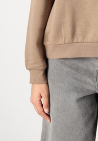 Sudadera beige con puños acanalados, combinada con pantalones gris claro. La tela parece suave y ligeramente texturizada, con un corte holgado.