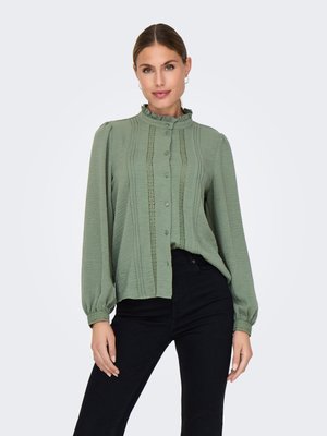 Mujer con una blusa de manga larga verde claro con cuello con volantes y pantalones negros, de pie frente a un fondo blanco liso.