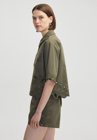 Chemise olive à col avec des manches courtes, ornée de dentelle décorative et d'accents en cristal le long de l'ourlet. Portée avec un short assorti.