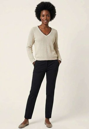 Femme aux cheveux naturellement bouclés, portant un pull beige à col en V, un pantalon noir et des ballerines claires, debout les mains dans les poches.