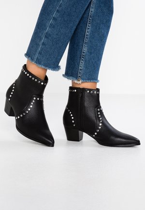 Zwarte leren enkellaarzen met zilveren studs, gedragen bij spijkerbroek met rafelige zoom, op een witte vloer.