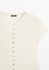 Kortemouw crèmegebreide cardigan met gestructureerd patroon en een verticale rij kleine ronde knopen aan de voorkant.