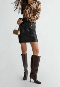 Langärmliges Oberteil mit braun-weißem Muster, schwarzer Minirock aus Leder mit Taschen und kniehohe, dunkelbraune Stiefel mit Krokodilprägung. Goldene Minihandtasche.