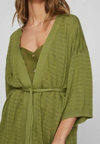 Cardigan en maille vert olive à ouverture frontale, manches larges et ceinture à nouer. Présente un motif texturé et est superposé à un top assorti.