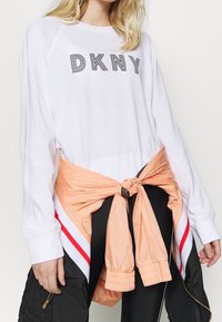Bílá košile s dlouhým rukávem a logem "DKNY". Růžová tkanina je uvázaná v pase přes černé kalhoty s červenými a bílými pruhy a elastickými manžetami.