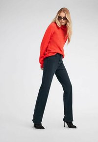 Maglione rosso a maglia grossa abbinato a jeans scuri. La figura si trova in una posa dinamica, indossando scarpe col tacco nere e occhiali da sole.