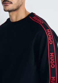 Svart hettegenser med røde "HUGO"-logoer langs skuldrene, laget av mykt materiale med ribbestrikket hals og avslappet passform.