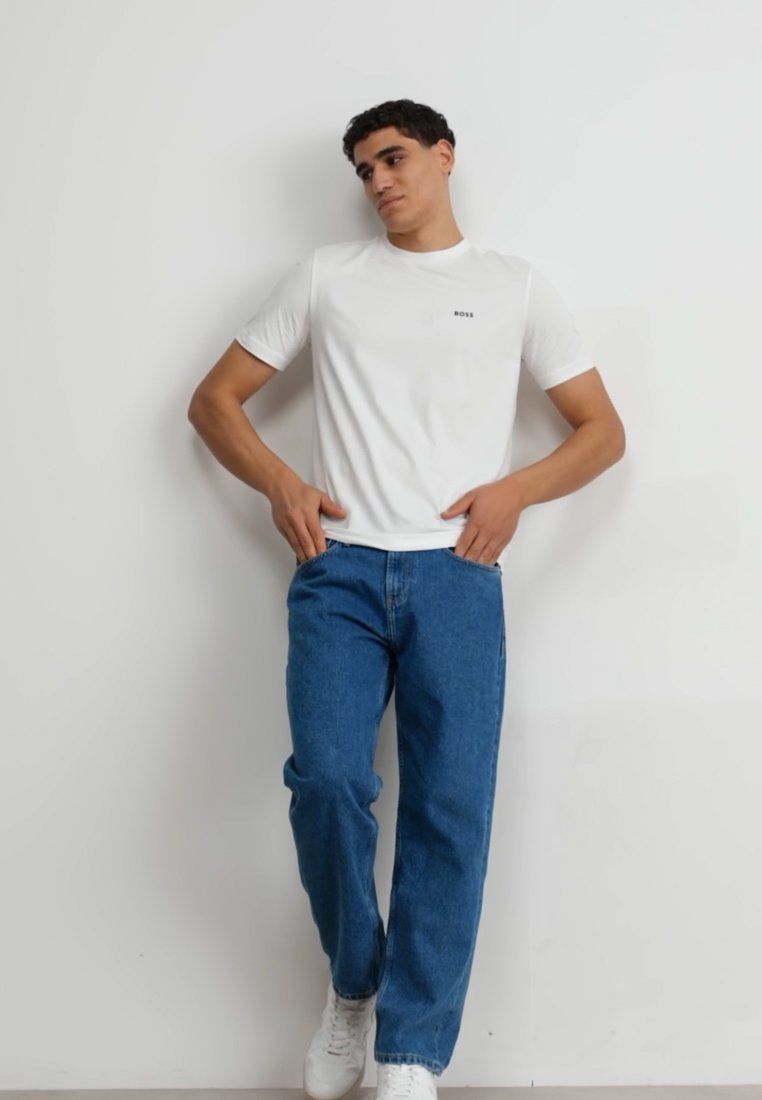 T-shirt blanc à col rond avec un petit logo noir "BOSS" sur la poitrine, associé à un jean en denim bleu et des baskets blanches.