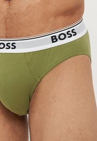 Zöld pamut bugyi fehér derékszíjjal, amelyen fekete "BOSS" felirat található. Az anyag sima és testhezálló fazonú.