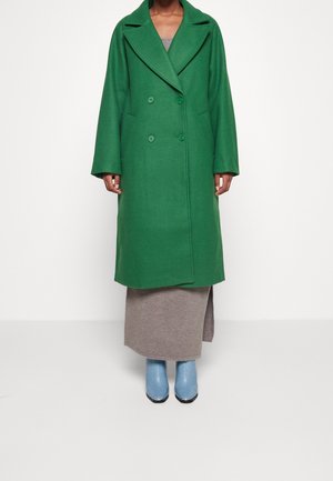 Femme portant un long manteau vert croisé sur une jupe beige avec une fente latérale et des bottines bleu clair, debout contre un mur blanc uni.