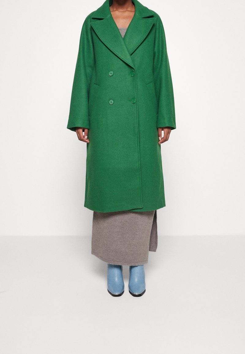 Femme portant un long manteau vert croisé sur une jupe beige avec une fente latérale et des bottines bleu clair, debout contre un mur blanc uni.