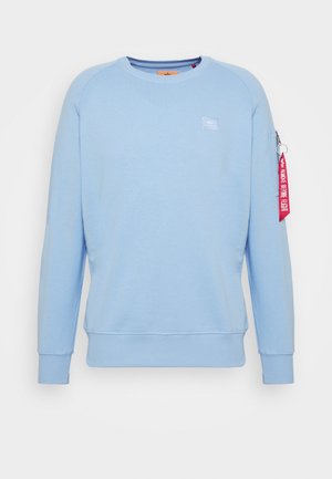 Lichtblauwe crewneck-trui met lange mouwen, geribbelde boorden en zoom, en een rood "Remove Before Flight"-label aan het ritszakje op de linker mouw.