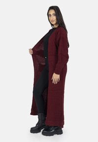 Cardigan lungo bordeaux con texture a coste e apertura frontale, abbinato a un top e pantaloni neri, indossato con stivaletti neri robusti.