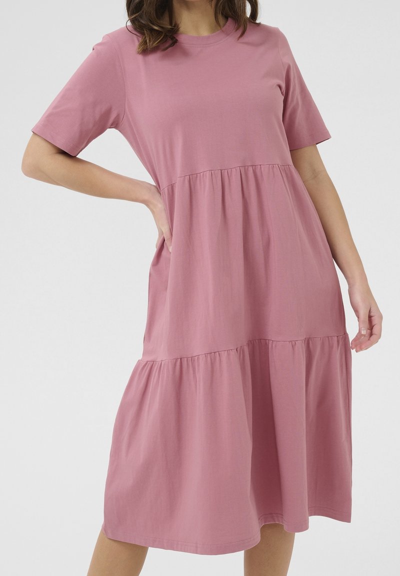 Robe en coton rose à manches courtes, présentant un design à volants et une coupe ample, se terminant juste en dessous du genou. Texture lisse.